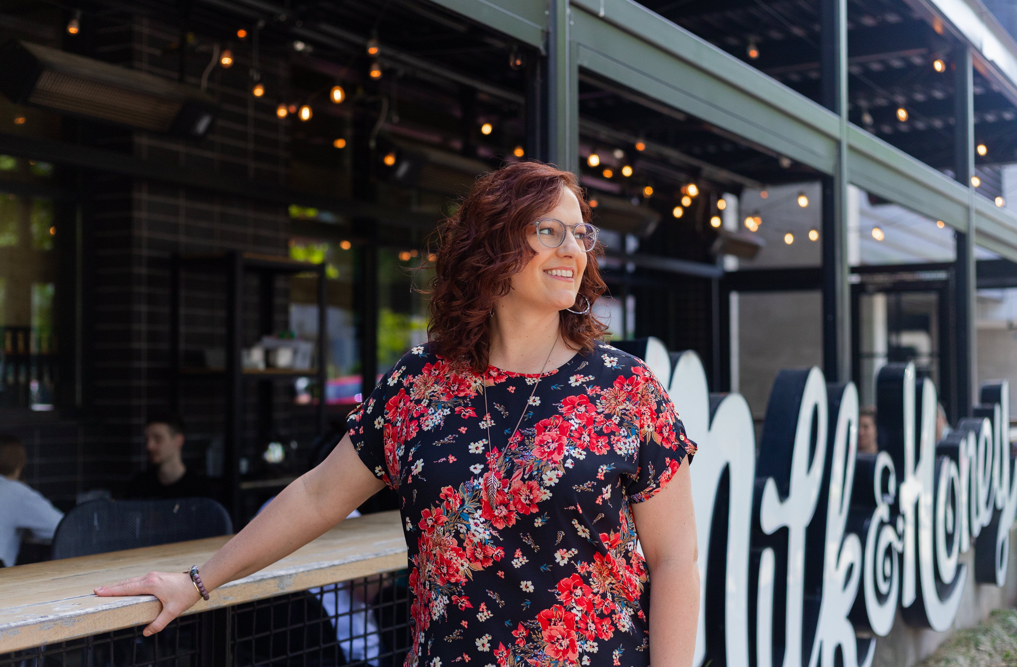 Woman in a floral shirt standing outside a building with 'Milk & Honey' on the wall.